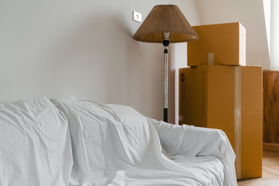 Vardagsrum under flytt med soffa täckt av vitt skynke, staplade flyttkartonger och en golvlampa